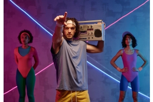 Ung idrottsman med svettband i pannan poserar med boombox medan två aerobiceskvinnor står i bakgrunden med 90-tals gymkläder