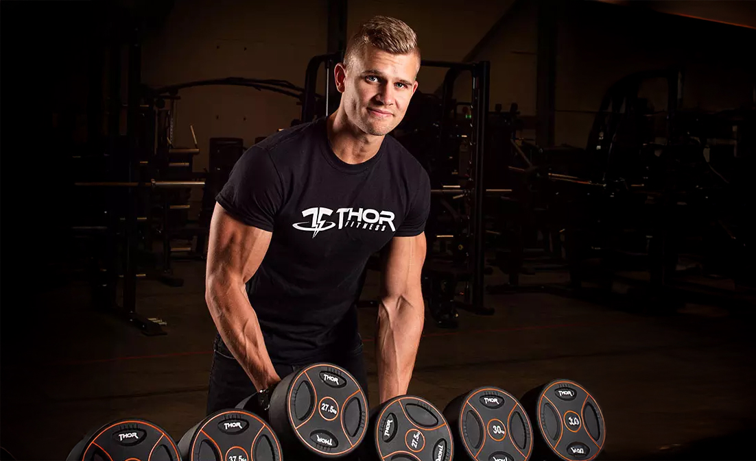 Viktor Rosengren står i ett mörkt gym och lyfter upp Thor Fitness hantlar